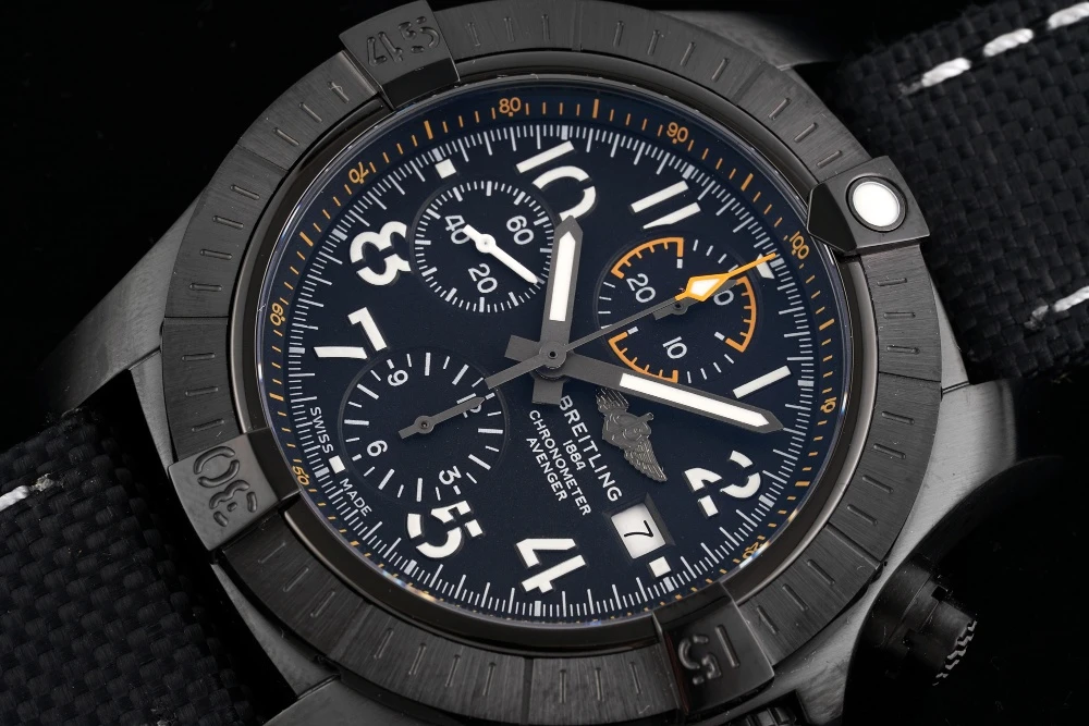 Close-up of Breitling Avenger Chronograph dark black dial with white Arabic numerals, orange-accented 30-minute counter, and date window at 3 o'clock